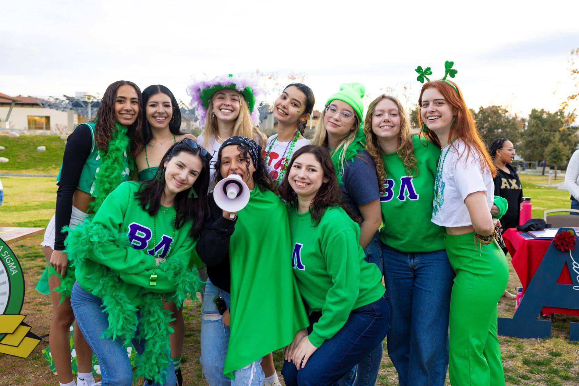 A group of students dressed in green pose together at an outdoor involvement fair event, some wearing festive accessories and sorority letters.