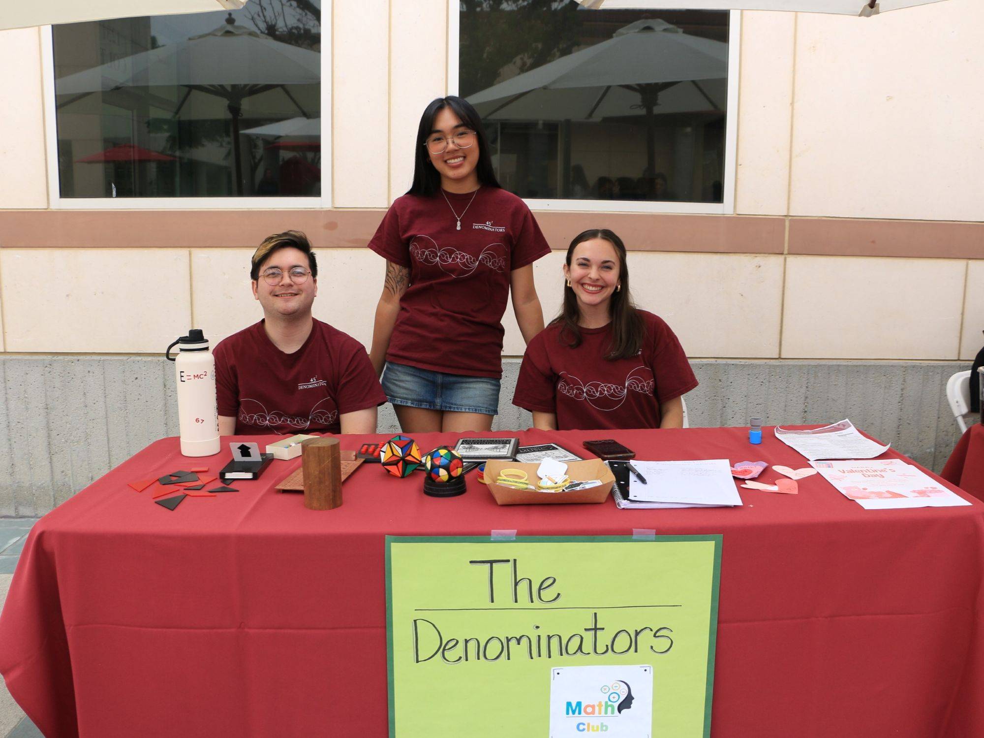 Three students stand behind a table labeled “The Denominators,