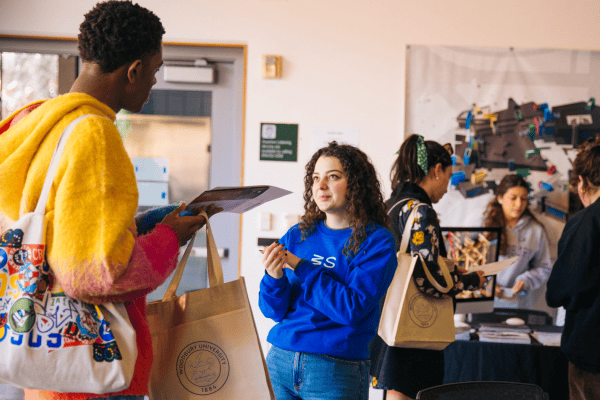 Two students speaking to each other, with more students grouped behind them interacting.